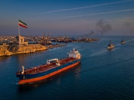 Oil tanker sailing through the Strait of Hormuz during US strikes on Iran, with Iranian flag flying near Bandar Abbas and U.S. naval ships patrolling nearby