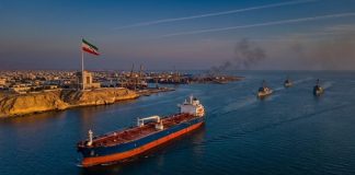 Oil tanker sailing through the Strait of Hormuz during US strikes on Iran, with Iranian flag flying near Bandar Abbas and U.S. naval ships patrolling nearby