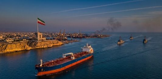 US Strikes on Iran: Self-Defense Strikes Target Missile Sites and Boats Amid Peace Talks Oil tanker sailing through the Strait of Hormuz during US strikes on Iran, with Iranian flag flying near Bandar Abbas and U.S. naval ships patrolling nearby